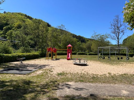 Spielplatz mit Rutsche, Schaukeln und Sandfläche, umgeben von Bäumen und Wiese.