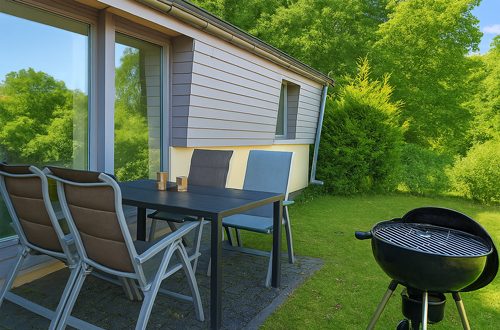 Terrasse vor dem Ferienhaus mit Tisch und Stühlen, Grill und Blick auf grünes Umland.