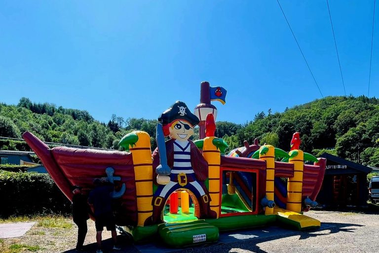 Kinderspielplatz Ferien-Campingpark Eifel Aufblasbares Piratenschiff mit bunten Figuren und Rutschen, umgeben von Natur.