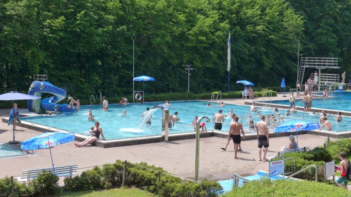 Badespaß im Freibad mit Menschen, Schwimmbecken und Sonnenschirmen in grüner Umgebung.