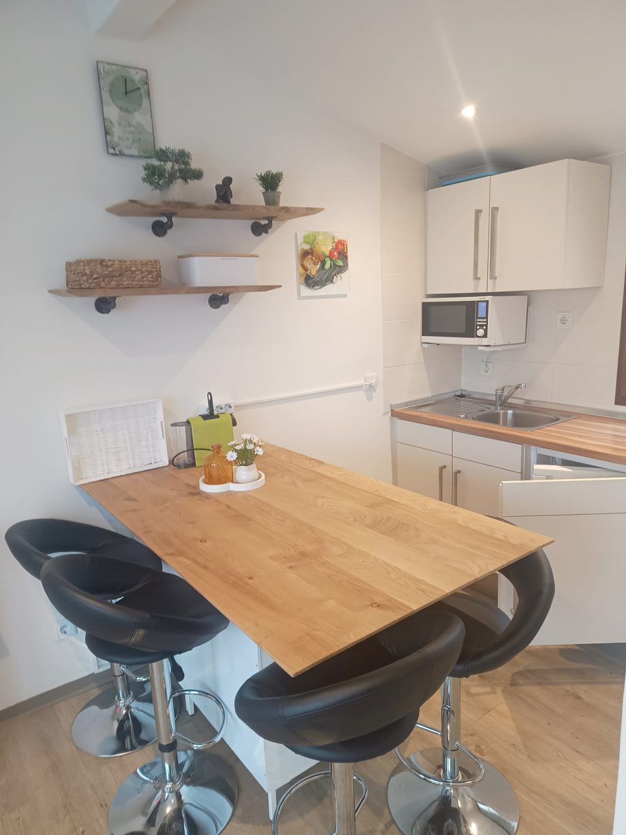 Blick in die Küche des Ferienhaus Moderne Küche mit Esstisch, schwarzem Stühlen und hängenden Regalen.