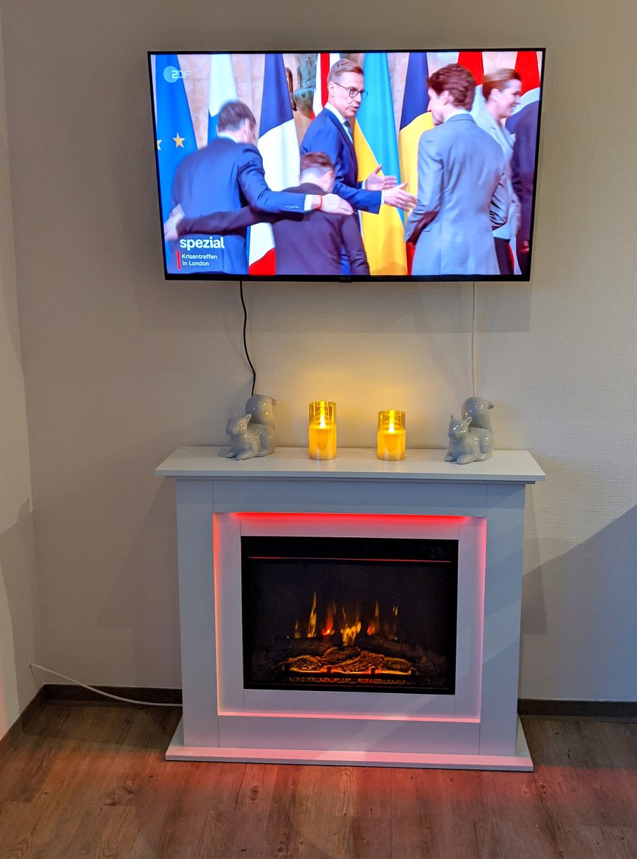 Elektrischer Kamin mit Beleuchtung Flackernder Kamin mit zwei Kerzen, Fernseher zeigt eine diplomatische Begegnung.