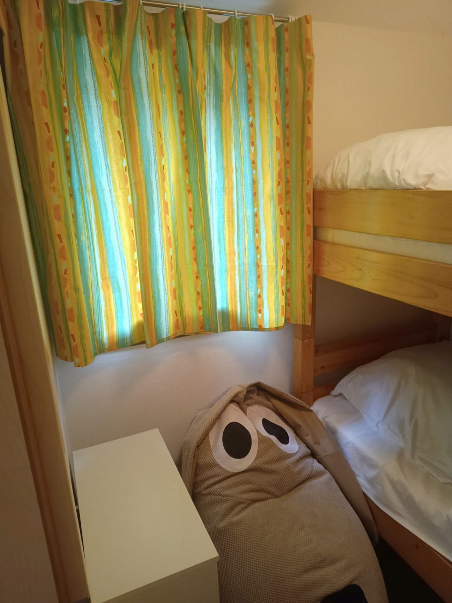 das 2. Schlafzimmer im Ferienhaus Bunte Vorhänge vor einem Fenster, ein Schrank und ein großes, rundes Kissen in einem Raum.