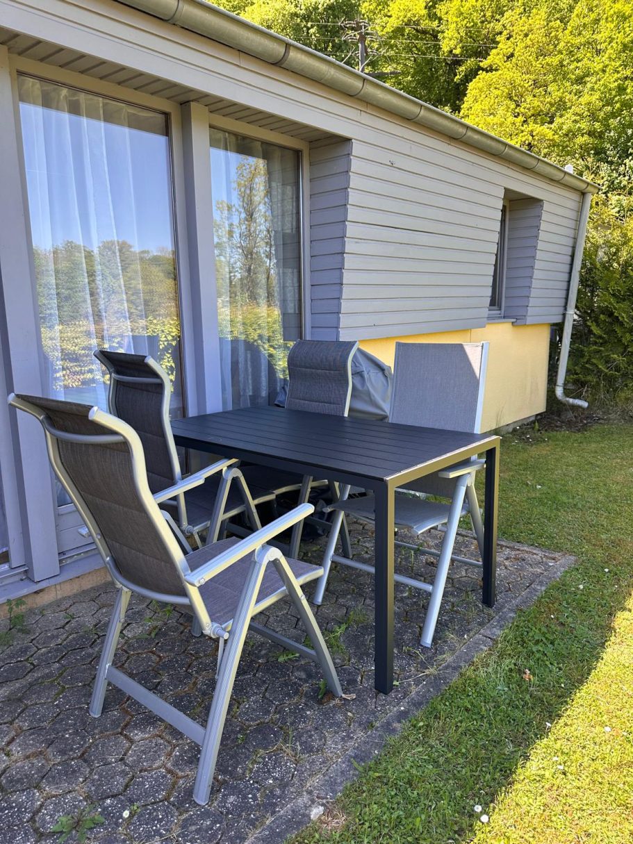 Blick auf die Terrasse vom Ferienhaus Ein Tisch mit Stühlen auf einer Terrasse, umgeben von grünem Rasen und Bäumen.