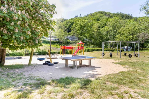 Spielplatz mit Rutsche, Schaukel und Sandbereich, umgeben von Bäumen und Wiese.