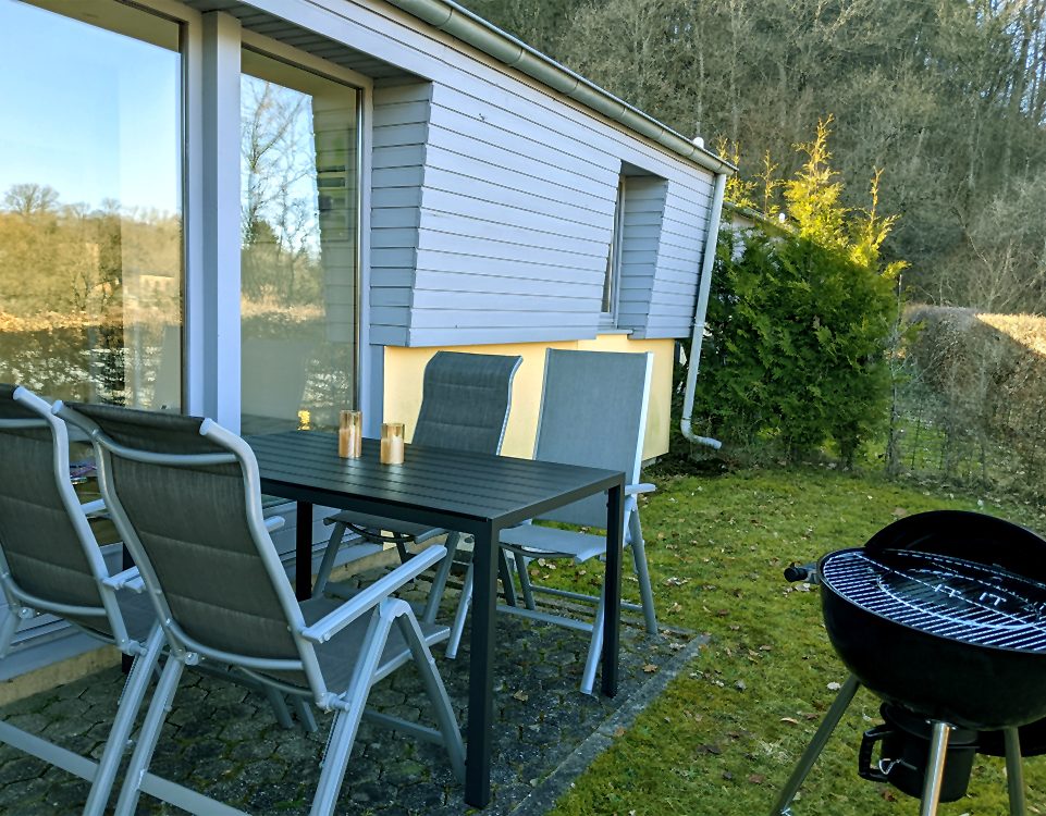 Terrasse mit Tisch, Stühlen und Grill vor dem Haus, umgeben von grüner Landschaft.
