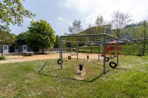 Spielplatz mit Schaukeln und Reifen, umgeben von Bäumen und Wiesen.