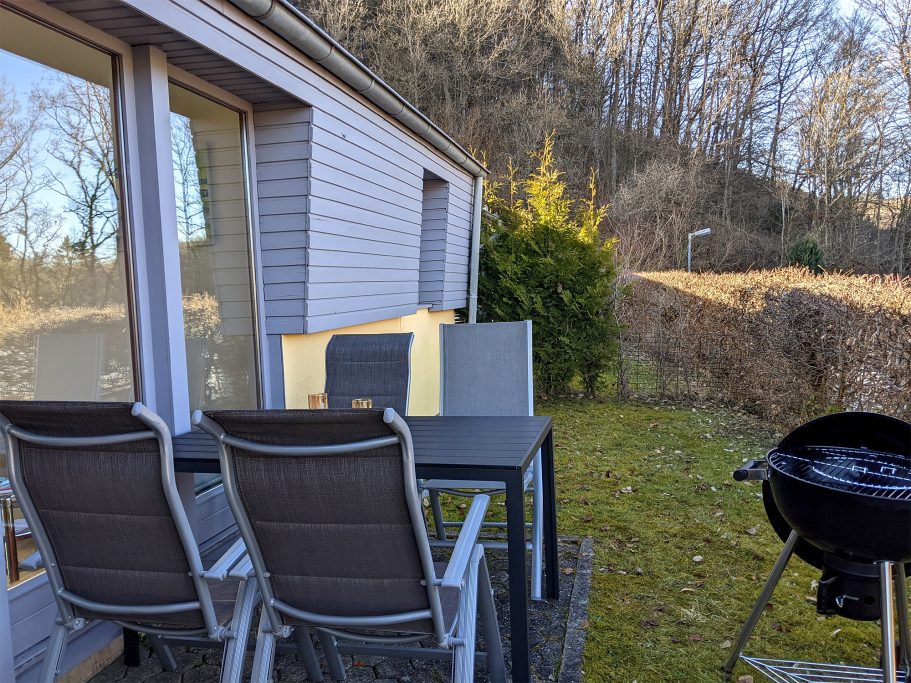 Die Terrasse mit Grill vom Ferienhaus Terrasse mit Tisch, Stühlen und einem Grill, umgeben von grüner Landschaft.