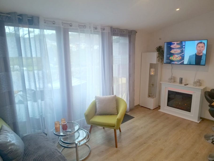 Blick ins Wohnzimmer vom Ferienhaus Heller Wohnraum mit Sofa, Tisch, Fernseher und großen Fenstern mit Vorhängen.